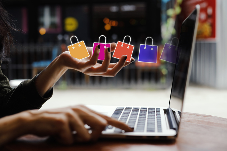 L’image montre une personne en train d’utiliser un ordinateur portable, une main sur le clavier et l’autre tendue vers l’avant, dans un geste interactif. Au-dessus de cette main sont superposées cinq icônes de sacs de shopping colorés e commerce