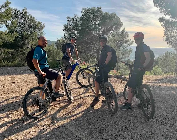 Quatre hommes posent avec leurs vélos tout-terrain dans un sentier boisé au coucher du soleil, portant des casques et des sacs à dos.