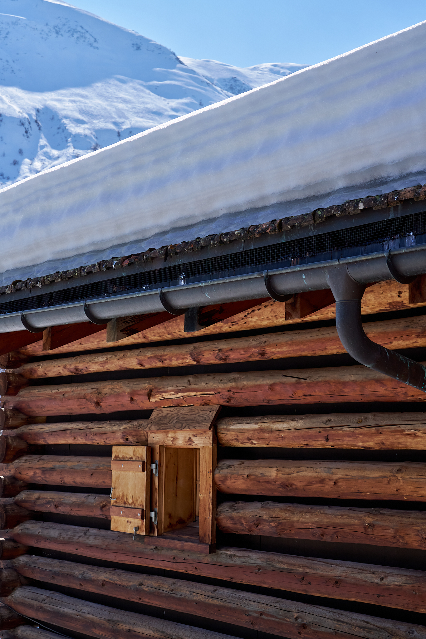 Holzhaus mit Schneeverwehungen und Dachrinnen vor verschneiten Bergen