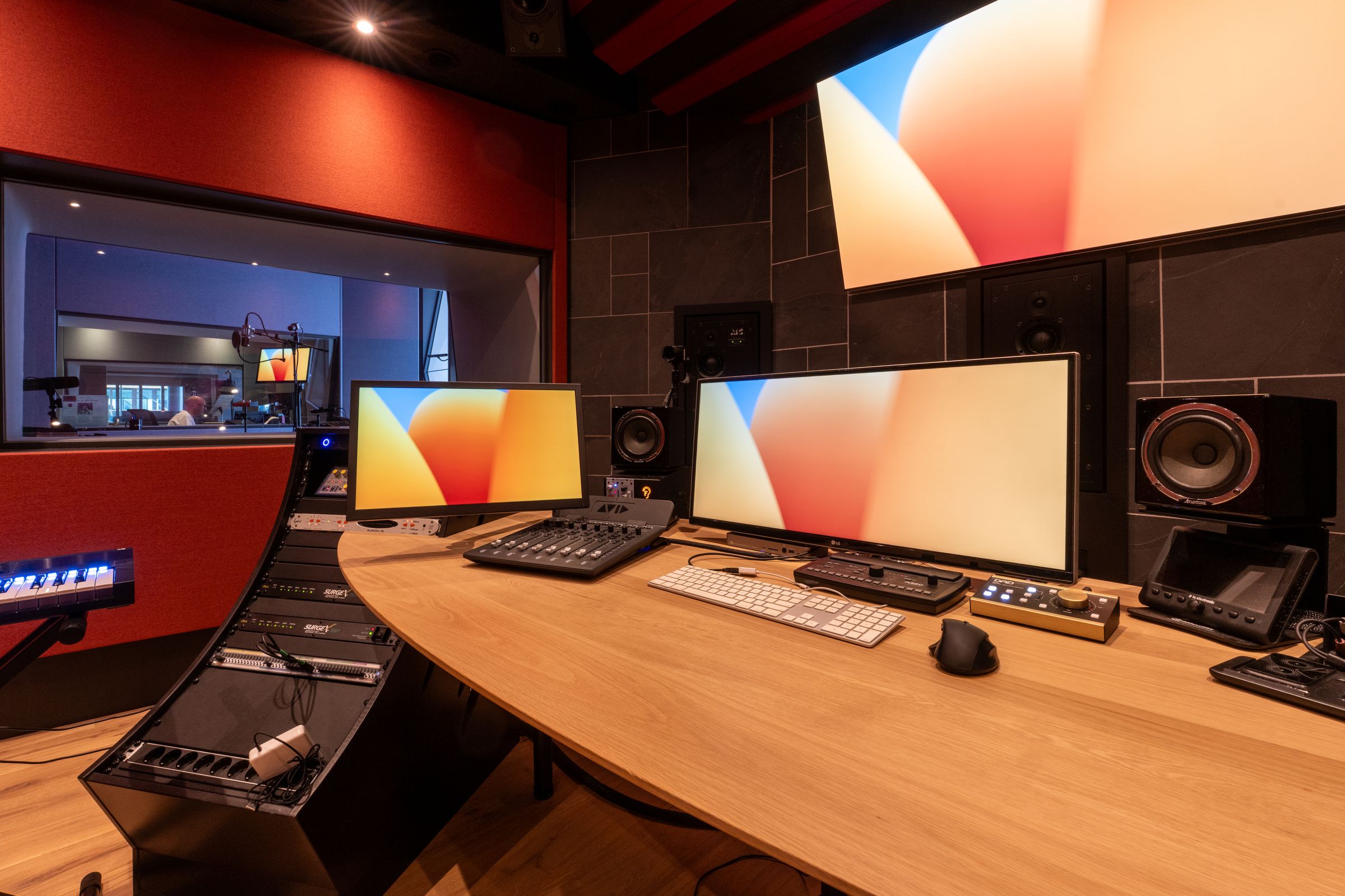 Werkplek in de rode studio van TFA met een breed scherm, toetsenbord en studiomonitoren