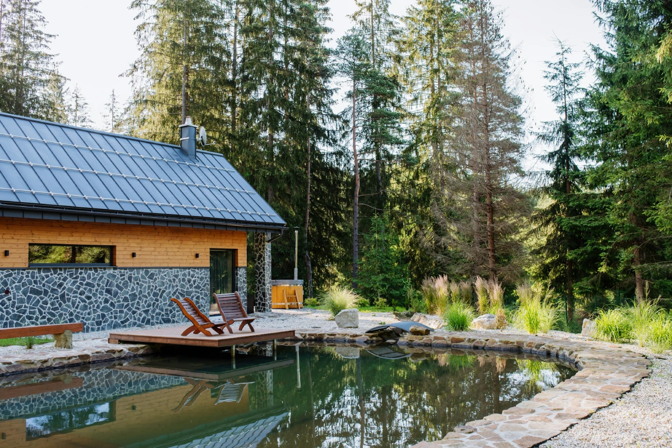 Cabin with stone and wood exterior beside a pond surrounded by trees and two wooden chairs on a small dock.