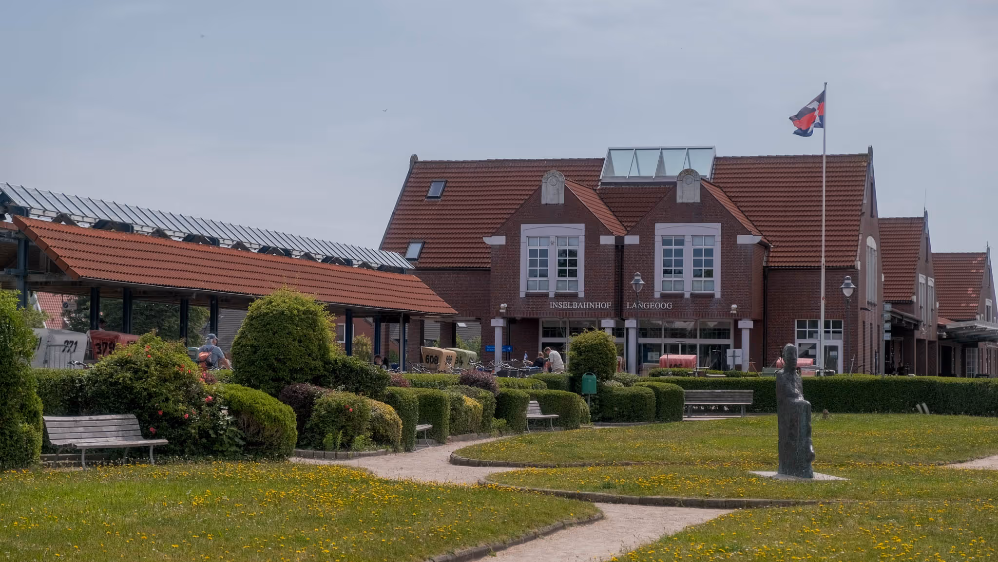 Bahnhof Inselbahnhof Langeoog mit roten Ziegeldächern, gepflegtem Garten, Sitzbänken und einer Statue im Vordergrund.