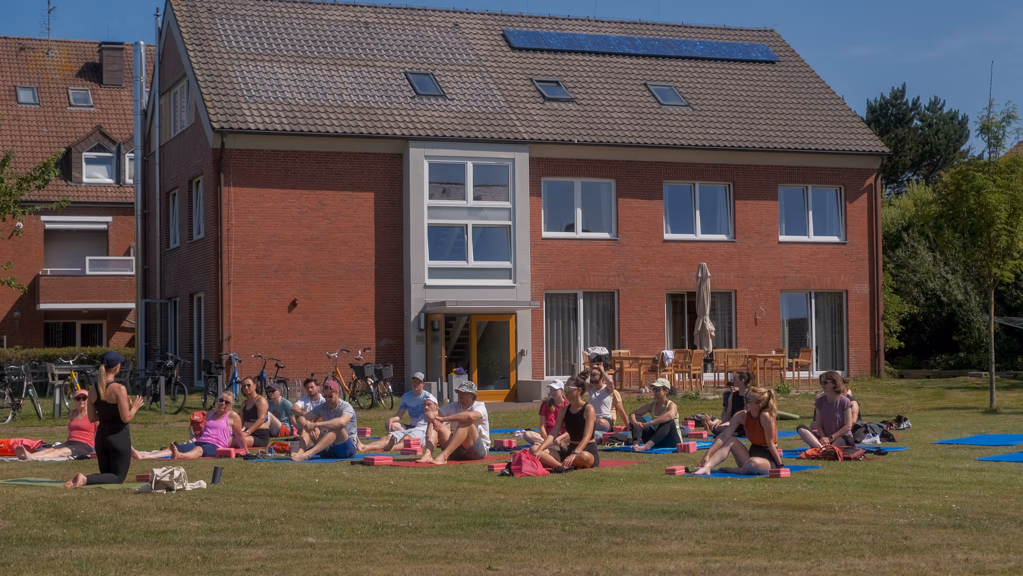 Gruppe von Menschen sitzt auf Yogamatten auf einer Wiese vor einem roten Backsteingebäude bei sonnigem Wetter.