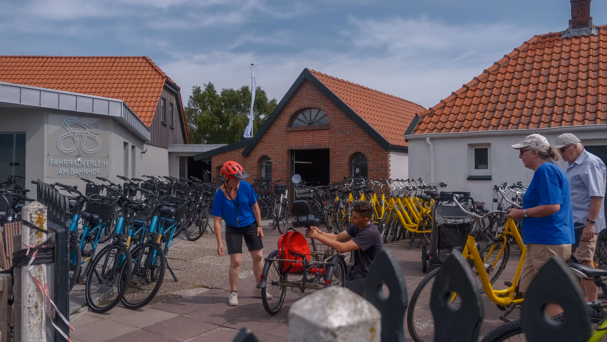 Fahrradverleih am Bahnhof mit mehreren schwarzen und gelben Fahrrädern, eine Frau mit rotem Helm steht neben einem Sitz-Dreirad, zwei ältere Personen mit gelben Fahrrädern.