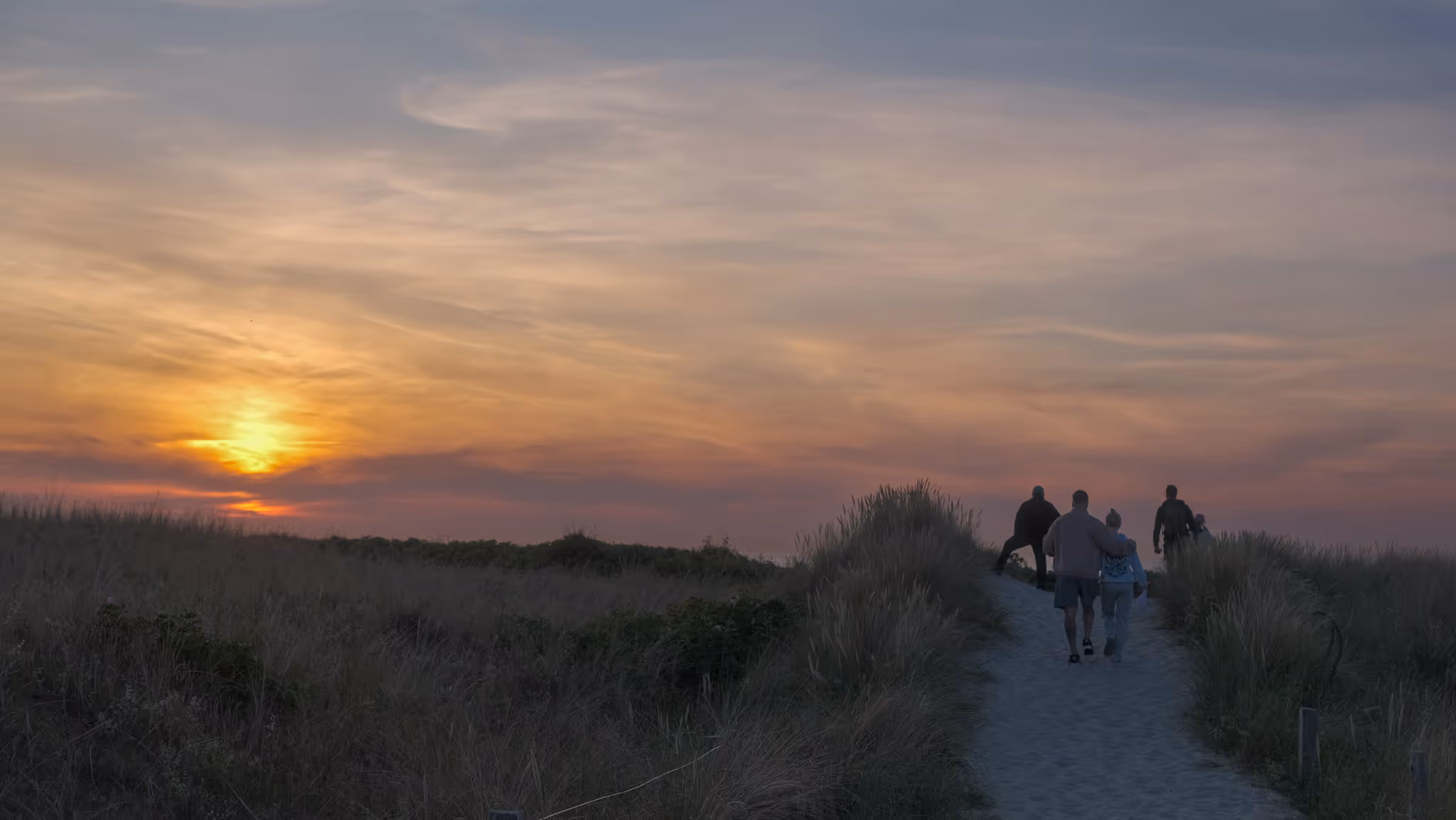 Gruppe von fünf Personen, die bei Sonnenuntergang über einen sandigen Pfad zwischen Dünen spazieren.