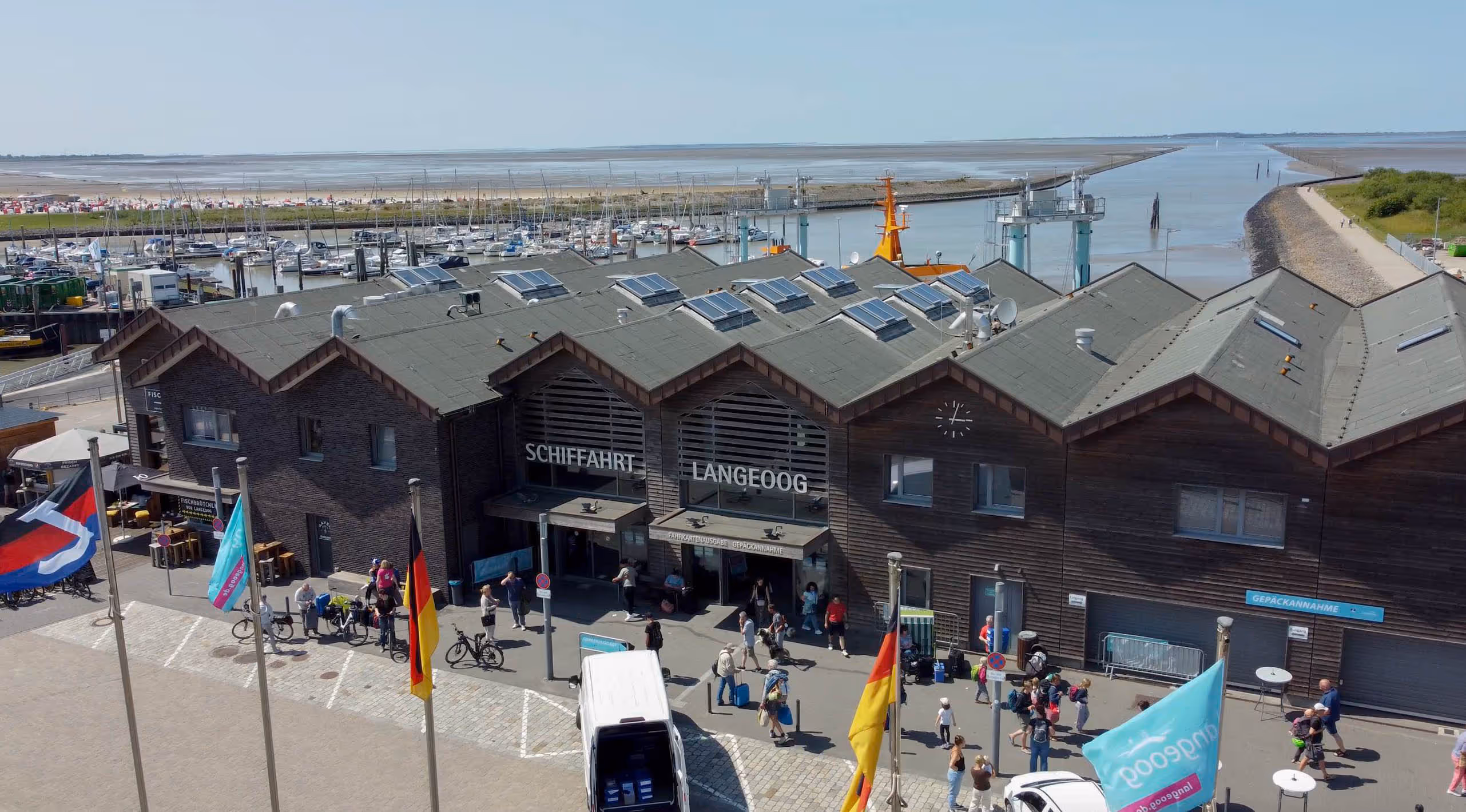 Menschen mit Koffern und Fahrrädern vor dem Gebäude "Schifffahrt Langeoog" am Hafen mit vielen Segelbooten im Hintergrund.
