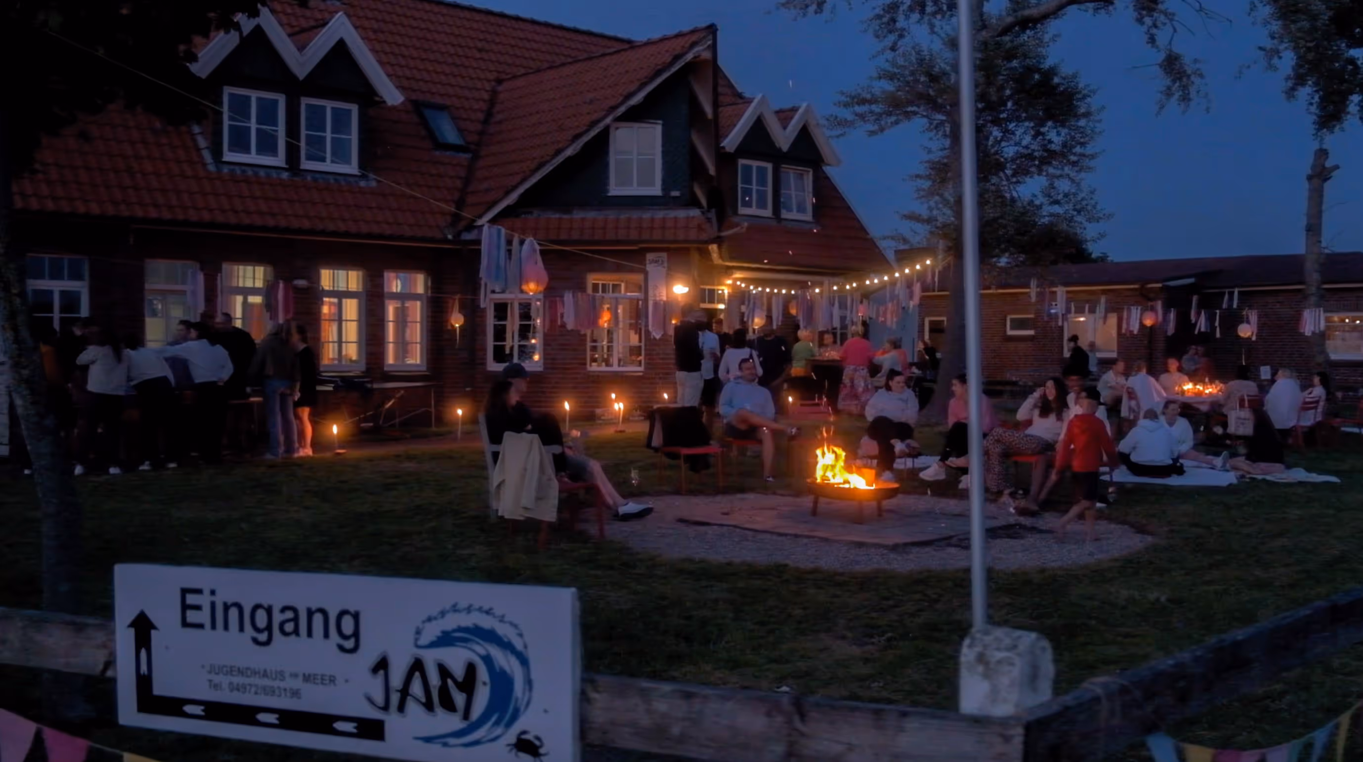 Abendliche Gartenfeier mit Menschen um ein Lagerfeuer vor einem Haus mit Beleuchtung und Girlanden.