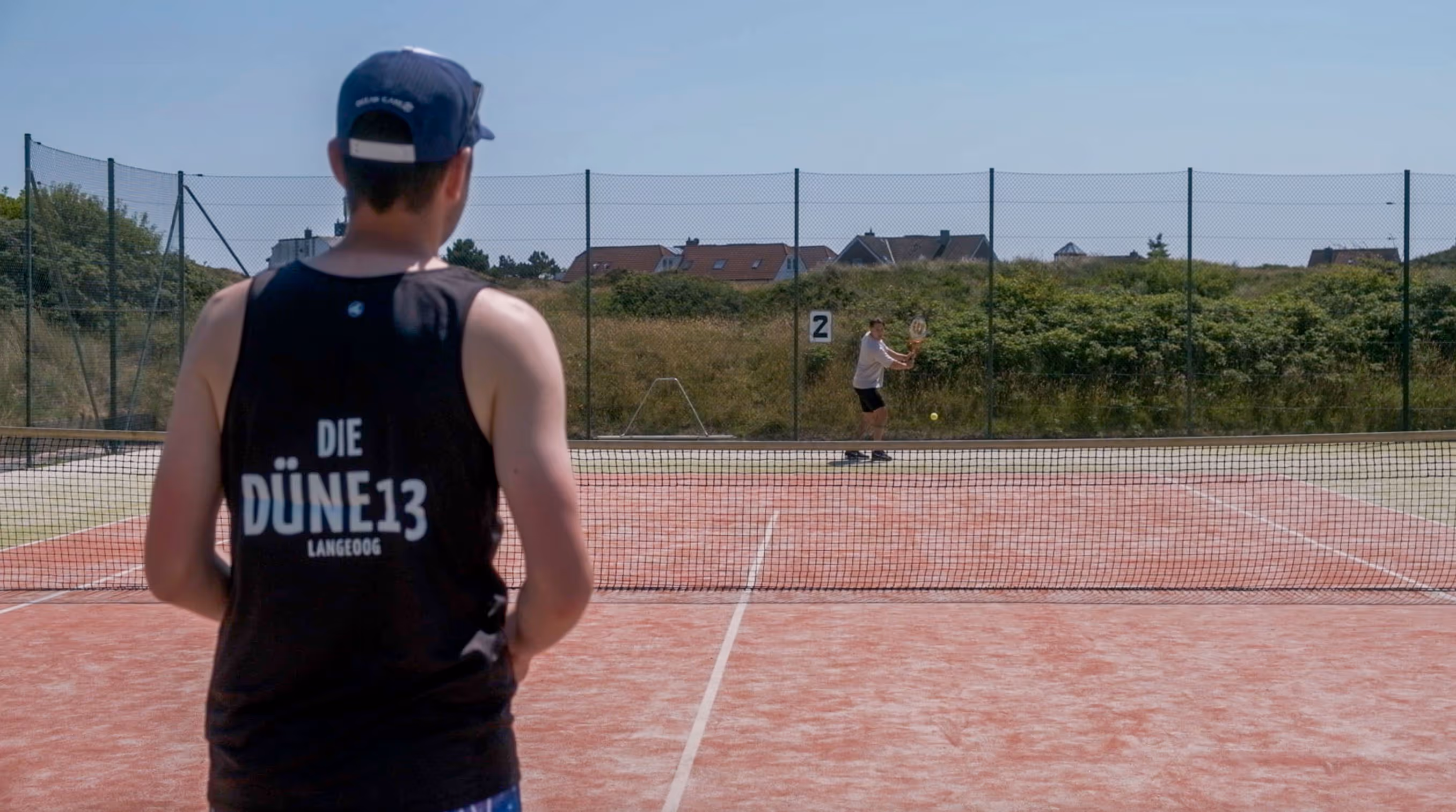Person in einem schwarzen Tanktop mit der Aufschrift ‘DIE DÜNE 13 LANGEOOG’ steht auf einem roten Tennisplatz, während ein anderer Spieler volley schlägt.