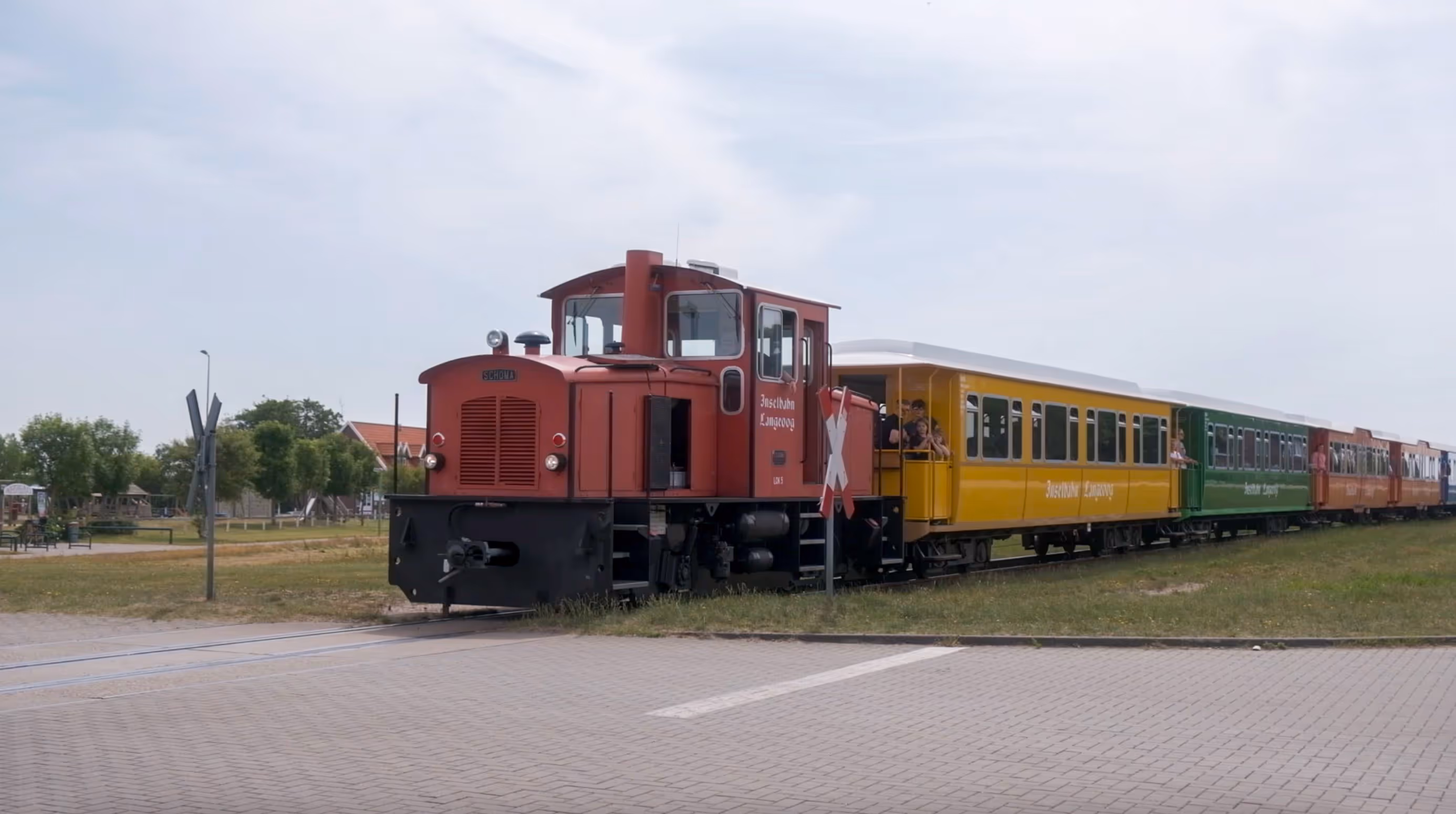 Rote Inselbahn mit bunten Waggons auf einer kurzen Eisenbahnstrecke nahe einem gepflasterten Bahnübergang.