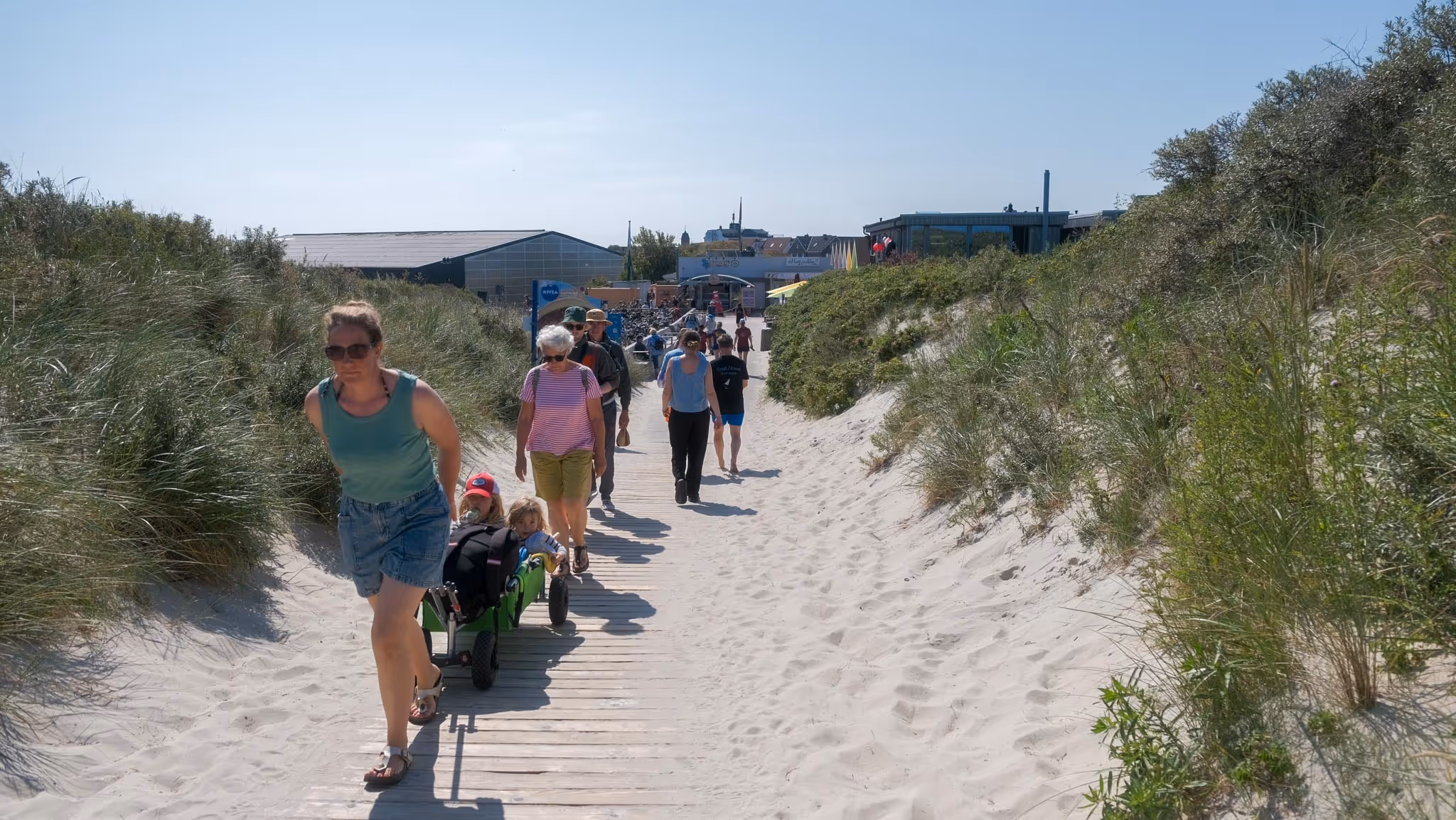 Menschen gehen auf einem Holzweg durch Dünen mit Sand und Gras an einem sonnigen Tag.