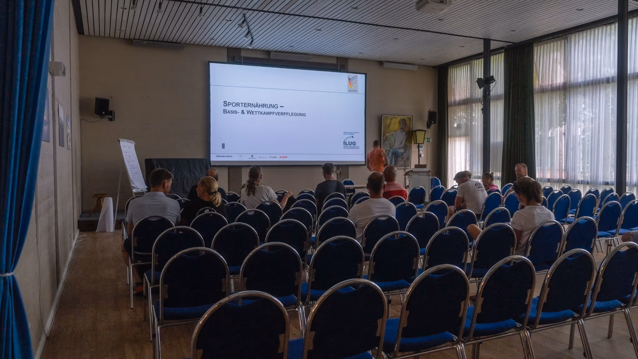 Menschen sitzen in einem Veranstaltungsraum mit blauen Stühlen vor einer Präsentationsleinwand zum Thema Sporternährung.
