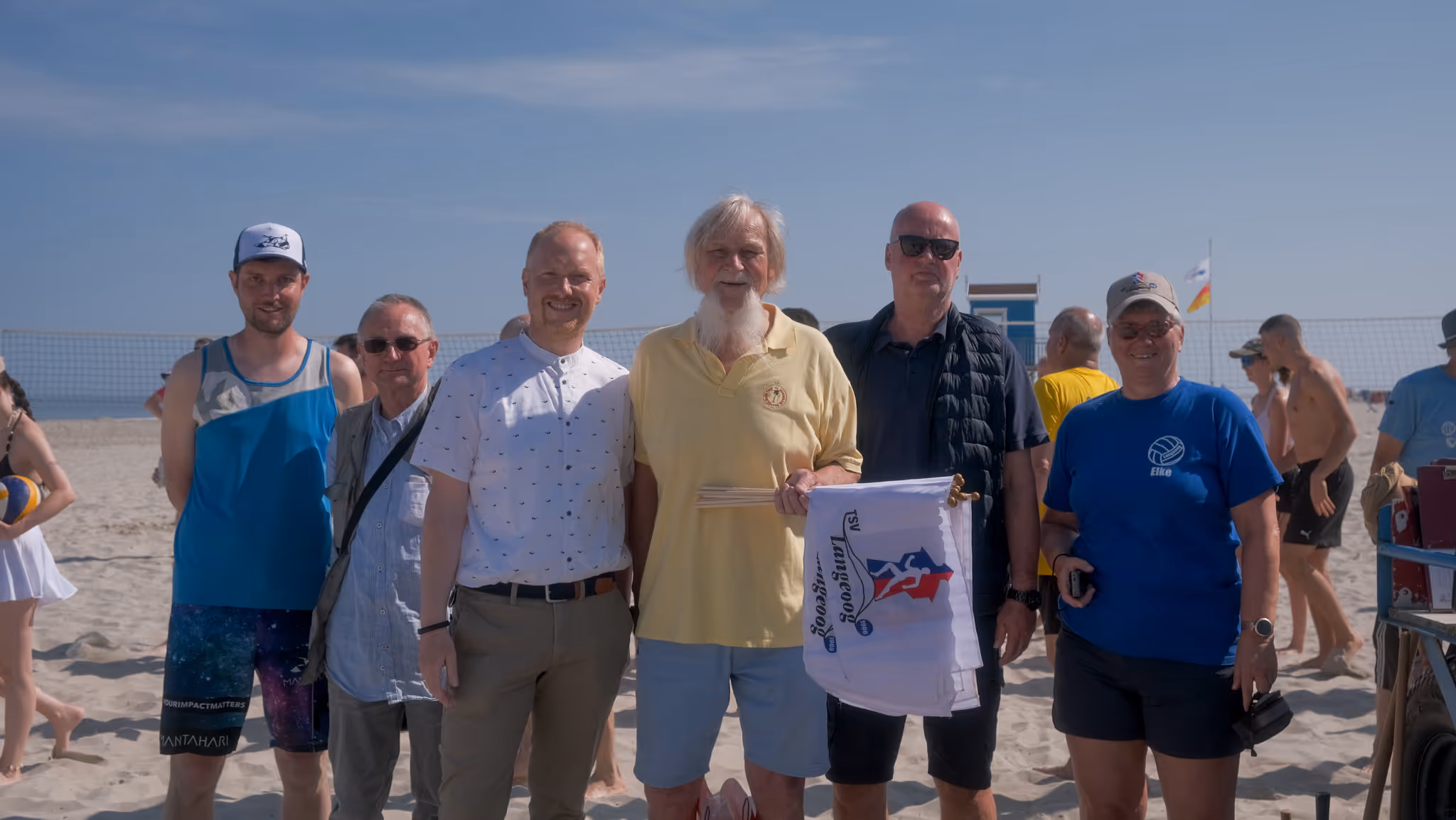Sechs Personen stehen an einem sonnigen Sandstrand vor einem Volleyballnetz, einer hält mehrere Flaggen mit Volleyball-Logo.