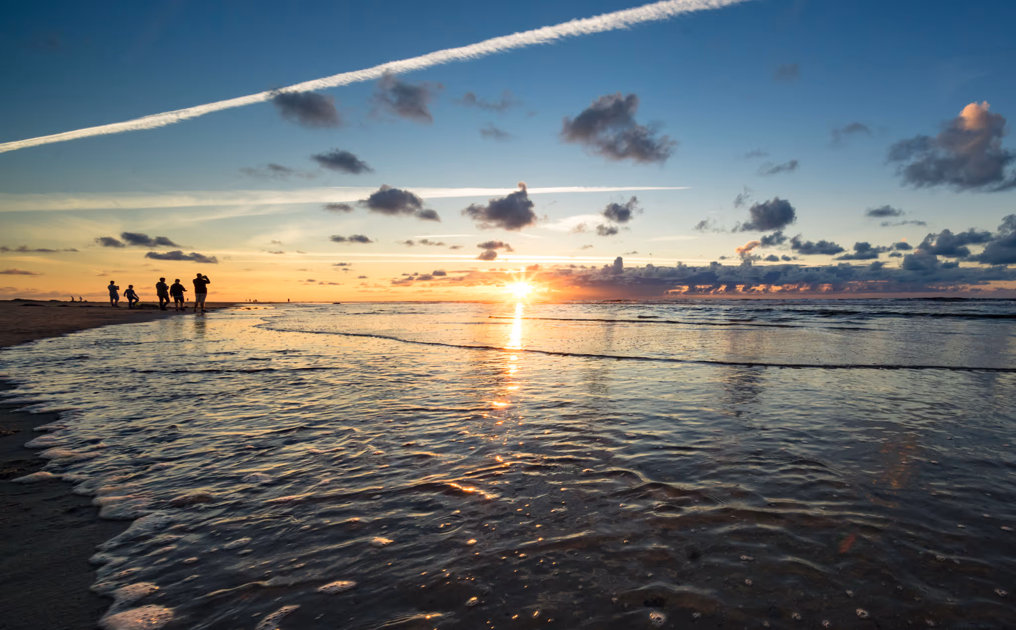 Sonnenuntergang über dem Meer mit Wellen am Strand und Silhouetten von Menschen am Ufer.