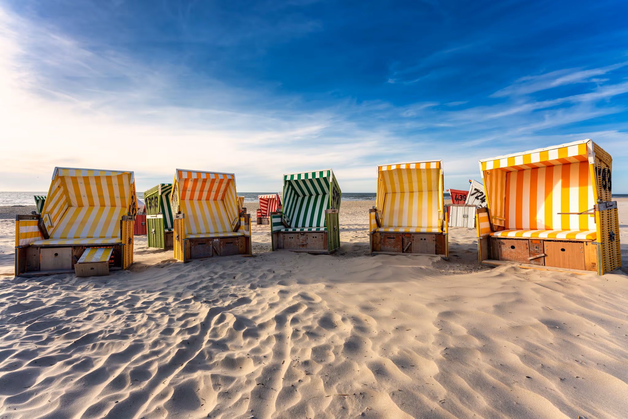 Mehrere bunte Strandkörbe mit gestreiftem Muster auf hellem Sandstrand unter blauem Himmel.