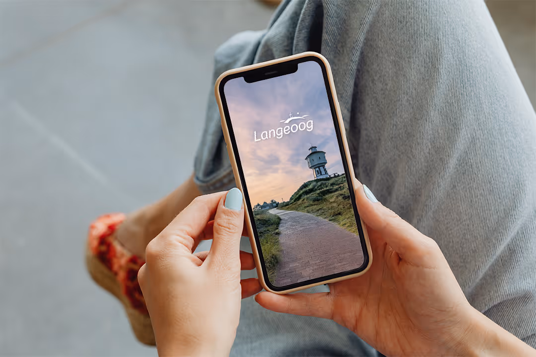 Eine Person hält ein Smartphone, auf dem eine Landschaft mit einem historischen Wasserturm und der Schrift 'Langeoog' angezeigt wird.