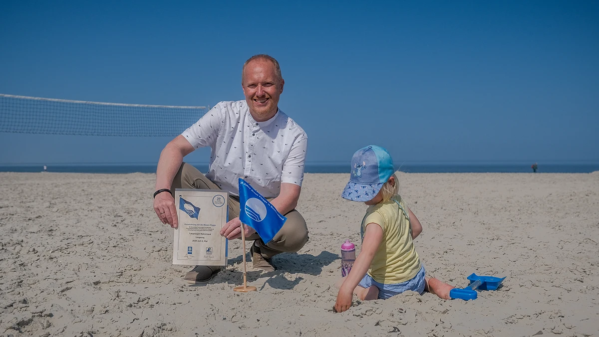 Mann kniet am Strand mit einer Auszeichnung und einer blauen Flagge, während ein Kind daneben im Sand spielt.