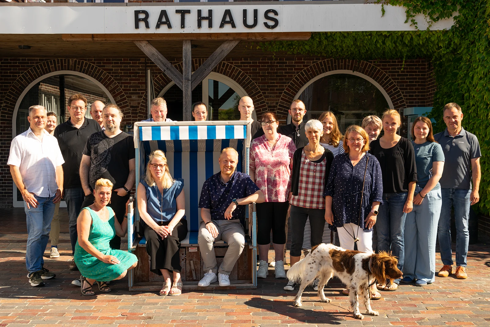 Gruppenfoto von 18 Personen der Gemeinde Langeoog vor dem Rathaus mit einem Hund und einer blau-weiß gestreiften Strandkorb in der Mitte.