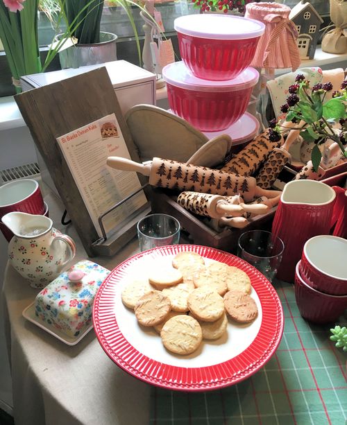 Plate of round cookies with tree imprints on a red-edged plate surrounded by decorative kitchenware including rolling pins with tree patterns, red containers, and floral ceramic items.