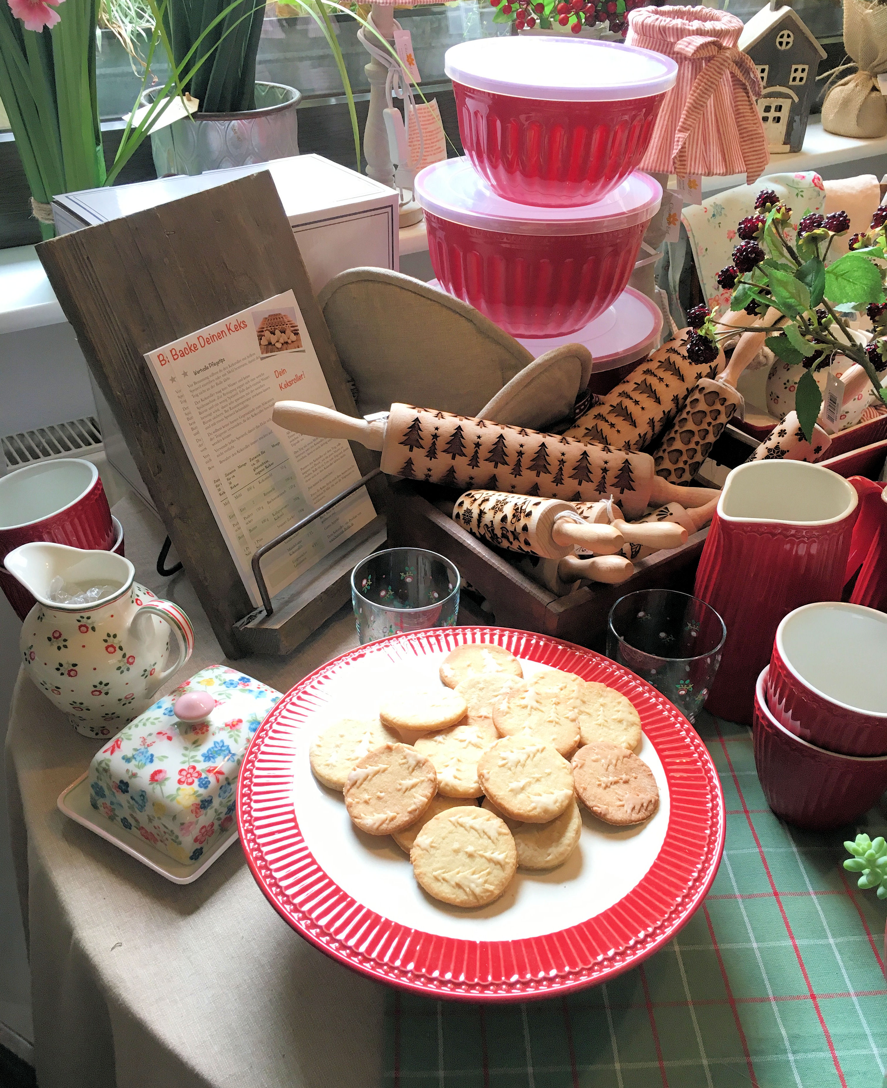Plate of round cookies with tree imprints on a red-edged plate surrounded by decorative kitchenware including rolling pins with tree patterns, red containers, and floral ceramic items.