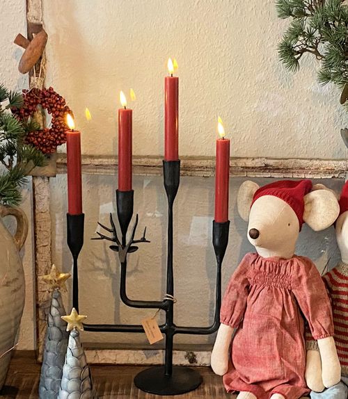Black metal candle holder with four lit red candles, decorated with a small metal reindeer head, next to a stuffed mouse doll in a red dress and hat.