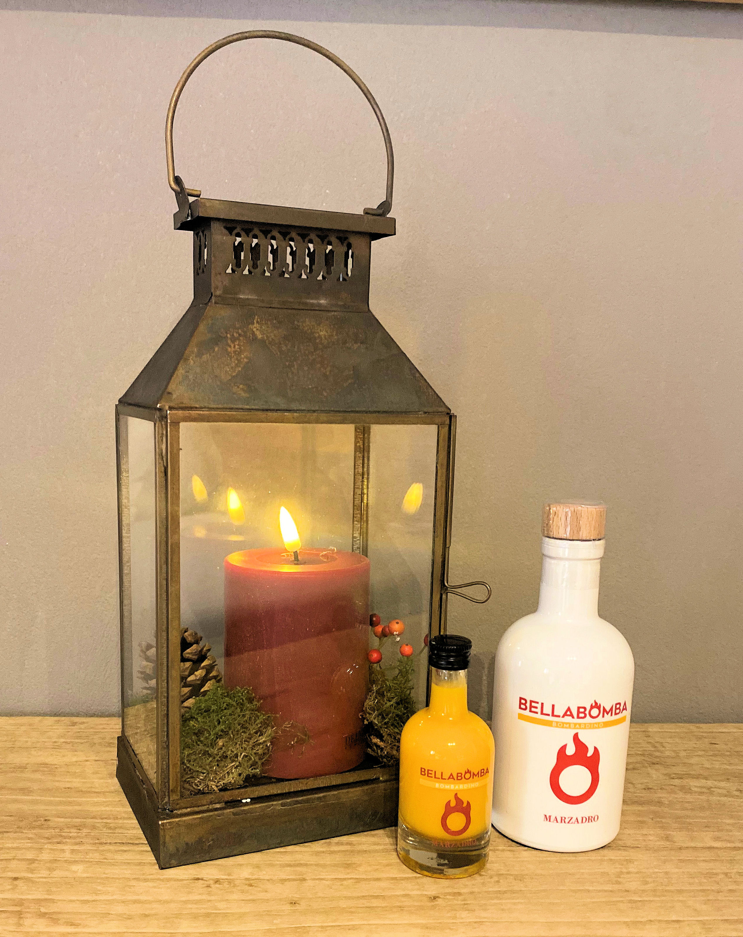 Rustic metal lantern with a lit red candle surrounded by pinecones and greenery on a wooden surface, next to two bottles of Bellabomba Bombardino liqueur.