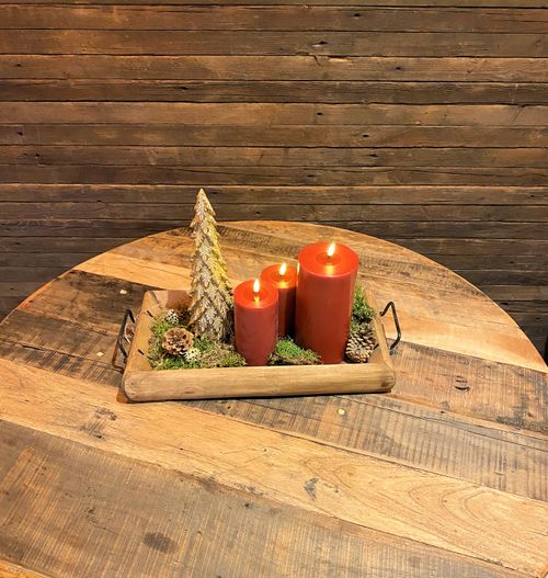 Wooden tray with three lit red candles, a decorative gold Christmas tree, pinecones, and green moss on a rustic wooden table.