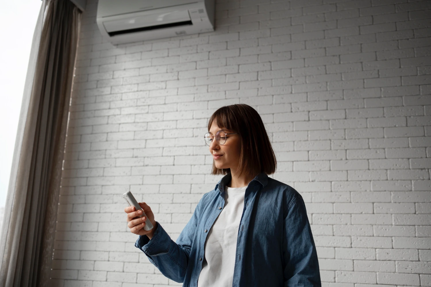 Person with glasses holding smartphone near air conditioner and brick wall