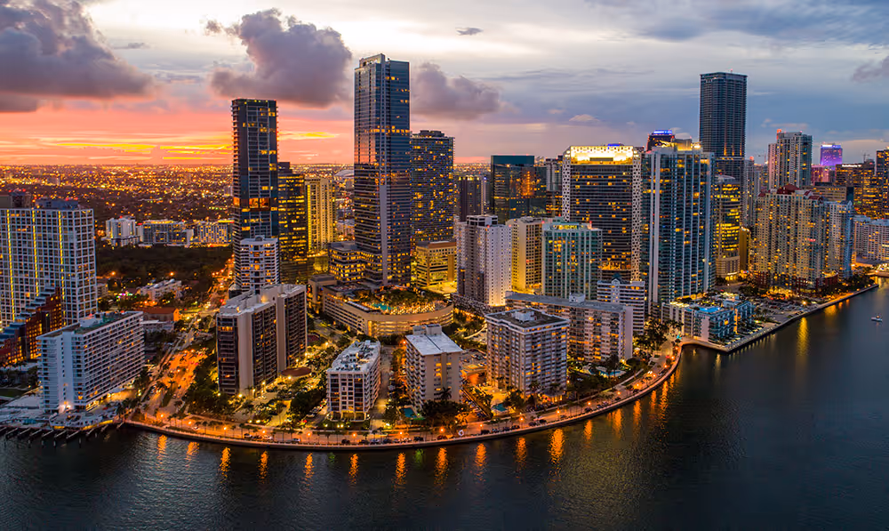 Tall buildings with illuminated windows line a waterfront at sunset, with clouds and city lights in the background.