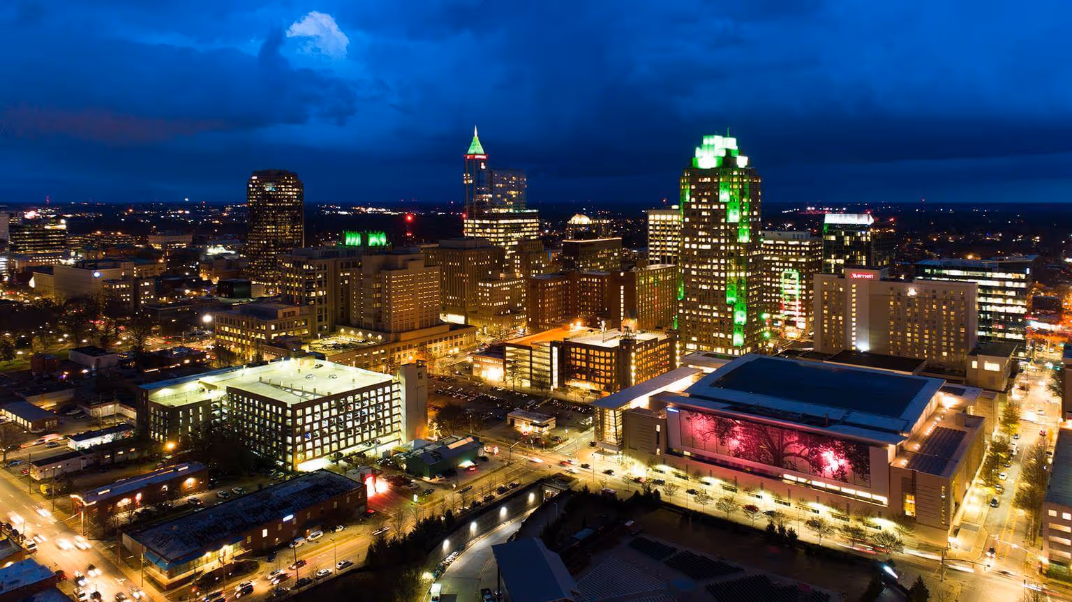 A cityscape at dusk with tall buildings illuminated by green and yellow lights under a dramatic cloudy sky.