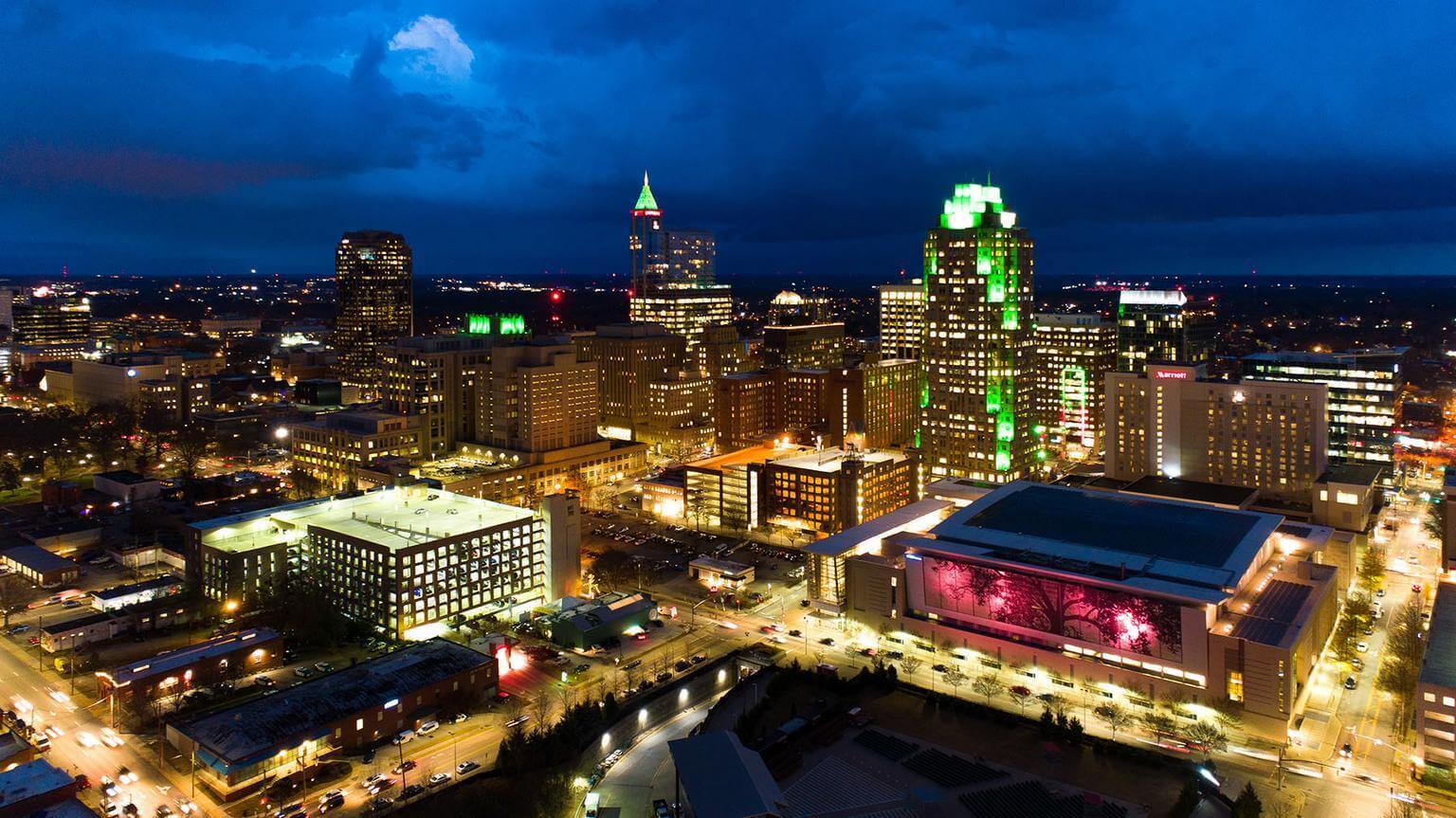 A cityscape at dusk with tall buildings illuminated by green and yellow lights under a dramatic cloudy sky.
