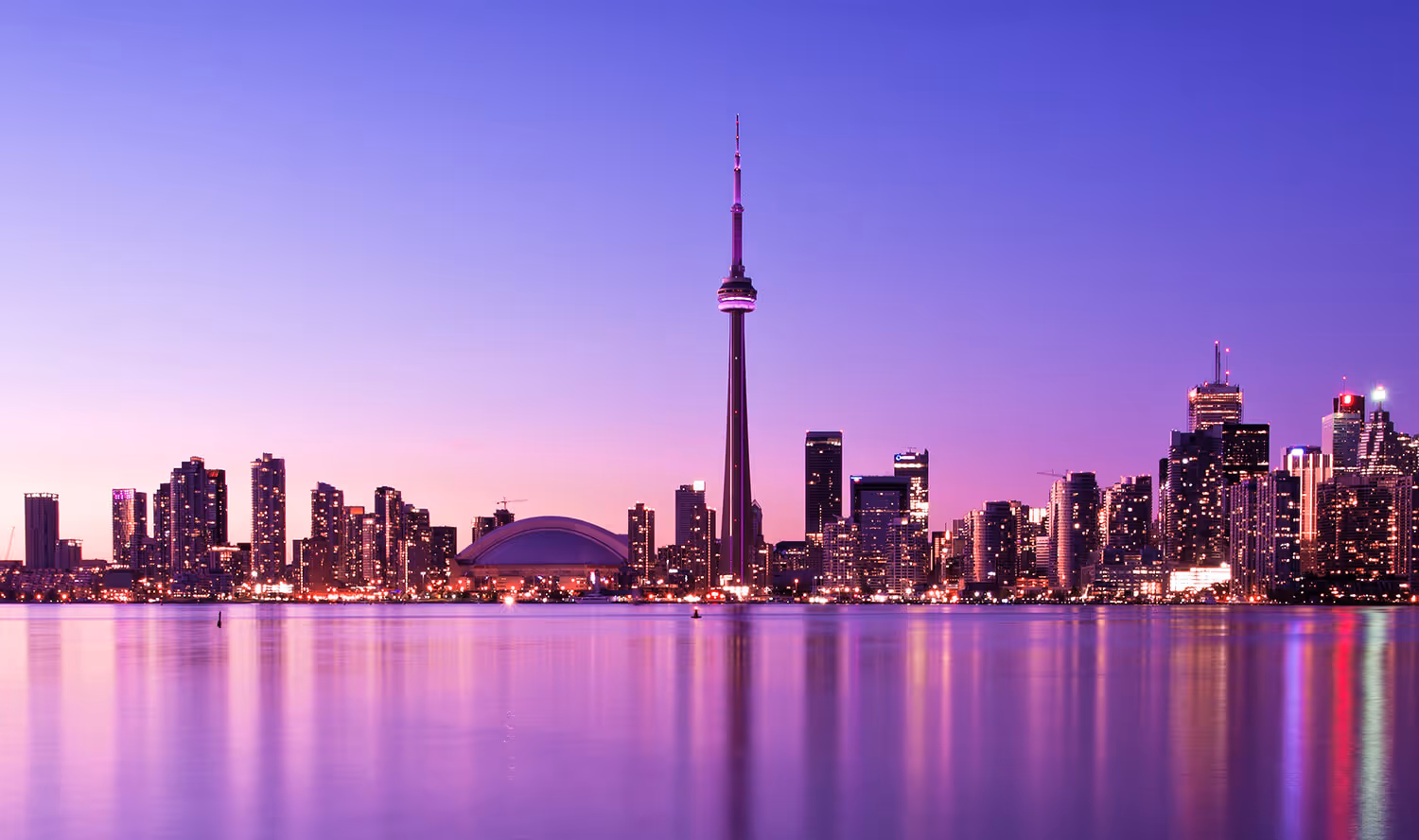 City skyline with illuminated buildings and a tall tower reflected on calm water at dusk with a purple sky.
