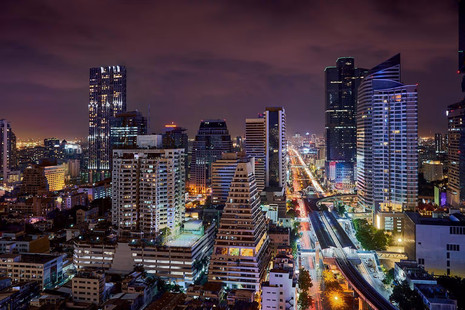 High-rise buildings with illuminated windows line a busy city street at night under a cloudy sky.