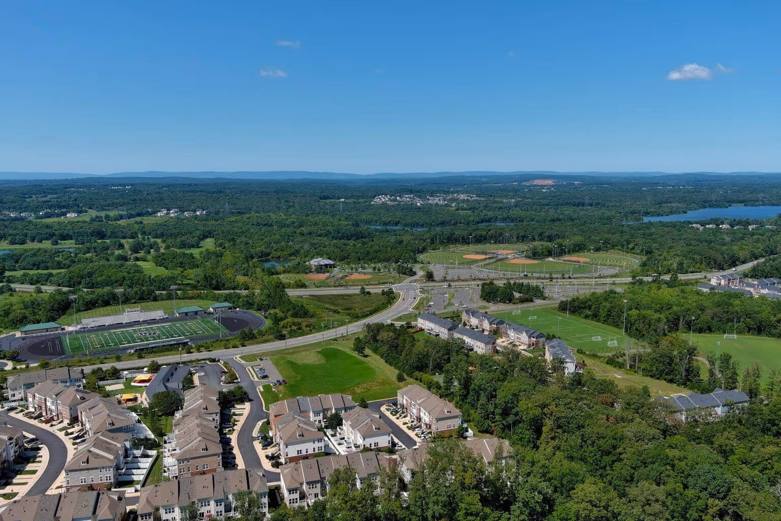 Aerial view of a suburban area with houses, sports fields, a stadium, roads, and surrounding green landscape.