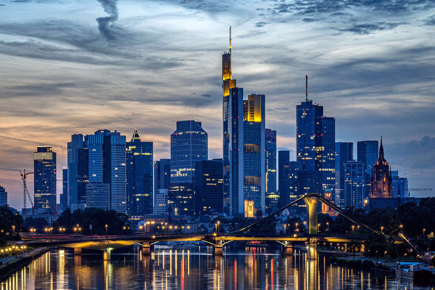 A city skyline with tall buildings, a bridge over a river, and illuminated windows at dusk.