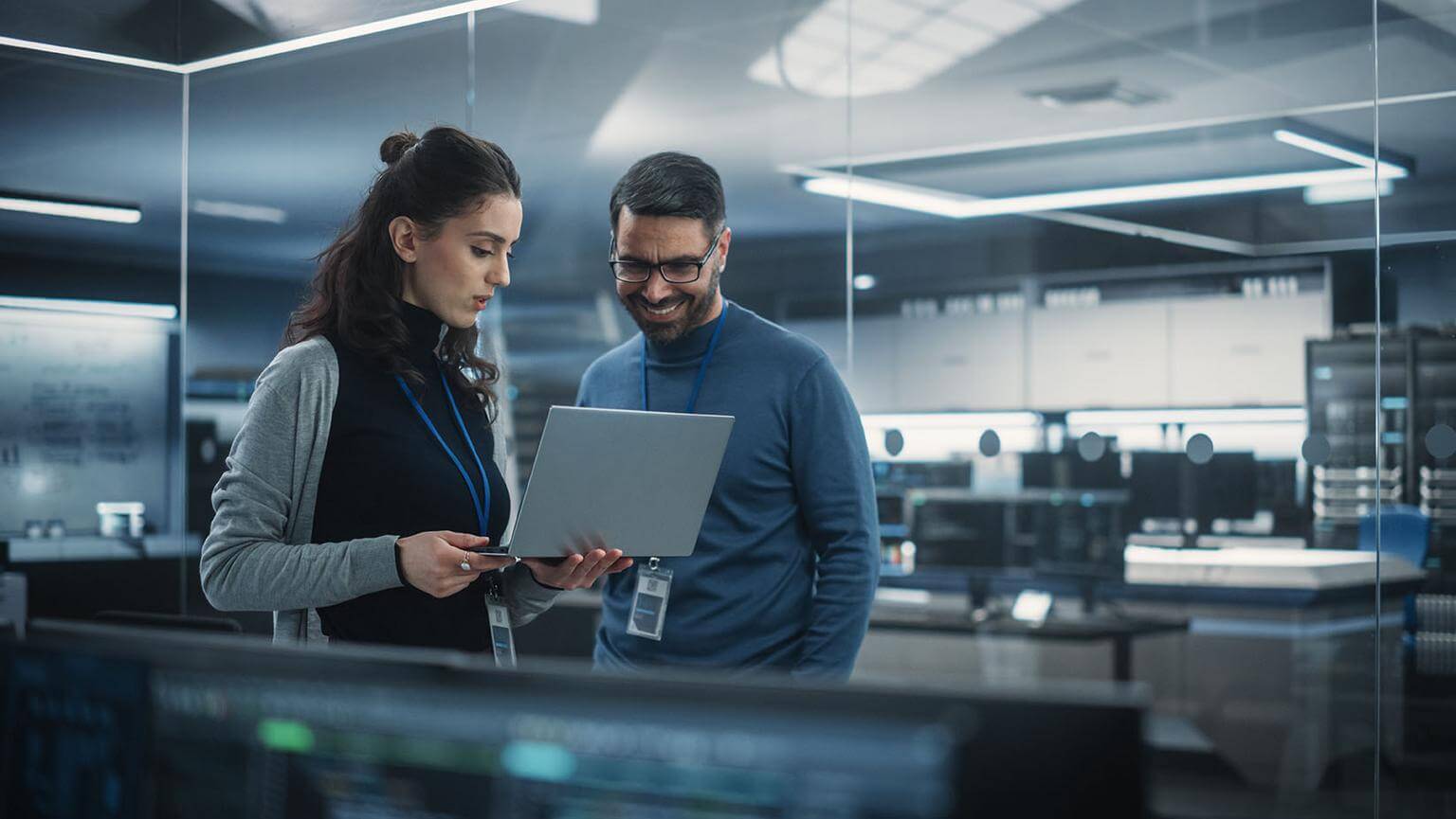 Two people wearing lanyards look at a laptop together in a modern office with computer equipment in the background.