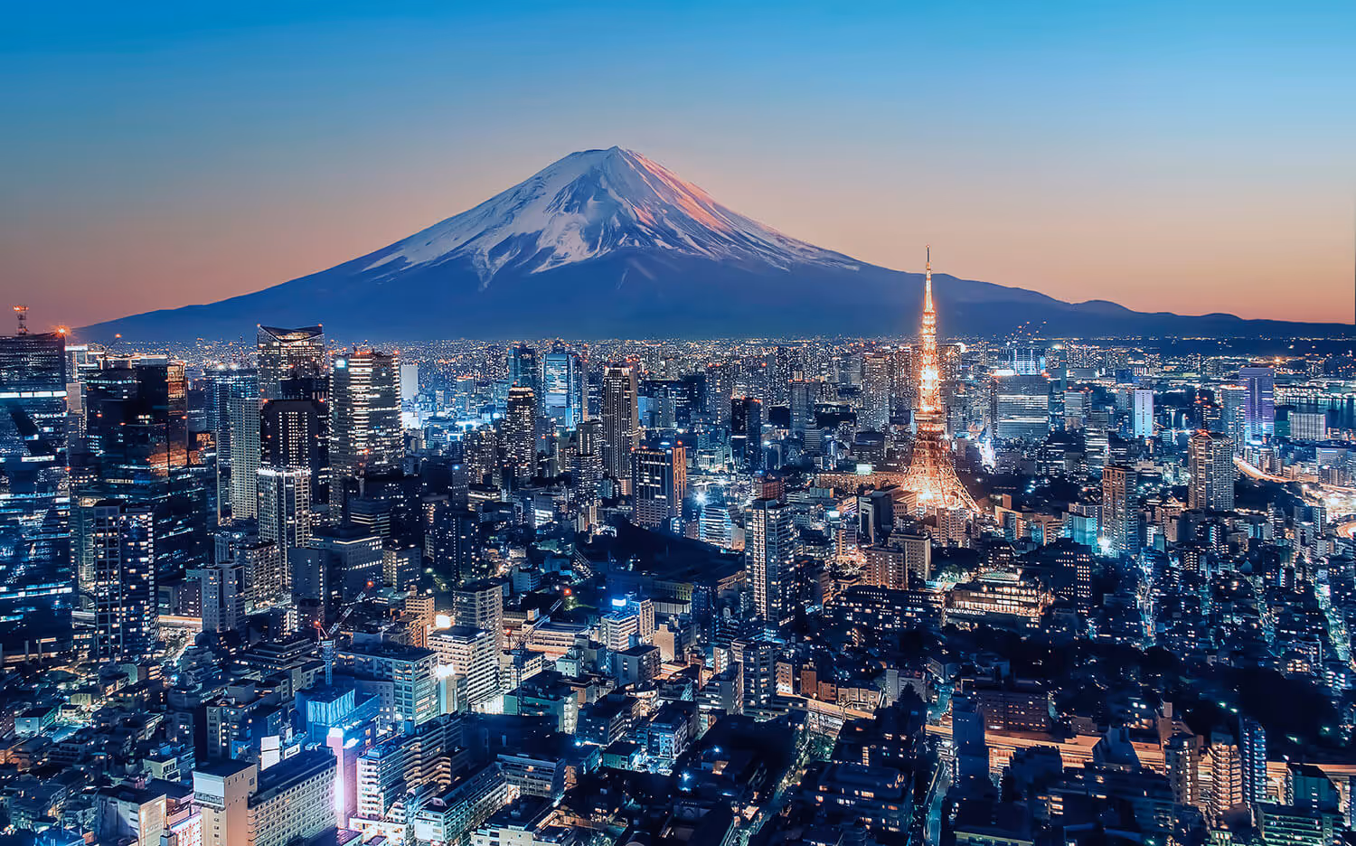 Tall buildings with many lights are seen at dusk with a snow-capped mountain and hills in the background.