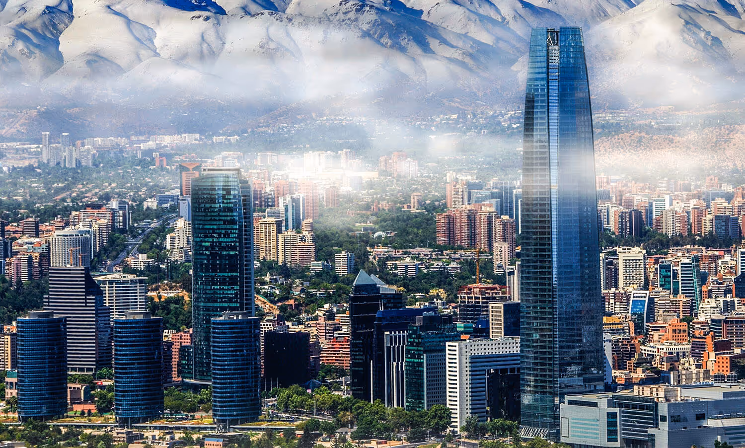 Tall glass skyscrapers and modern buildings with snow-covered mountains in the background under a partly cloudy sky.