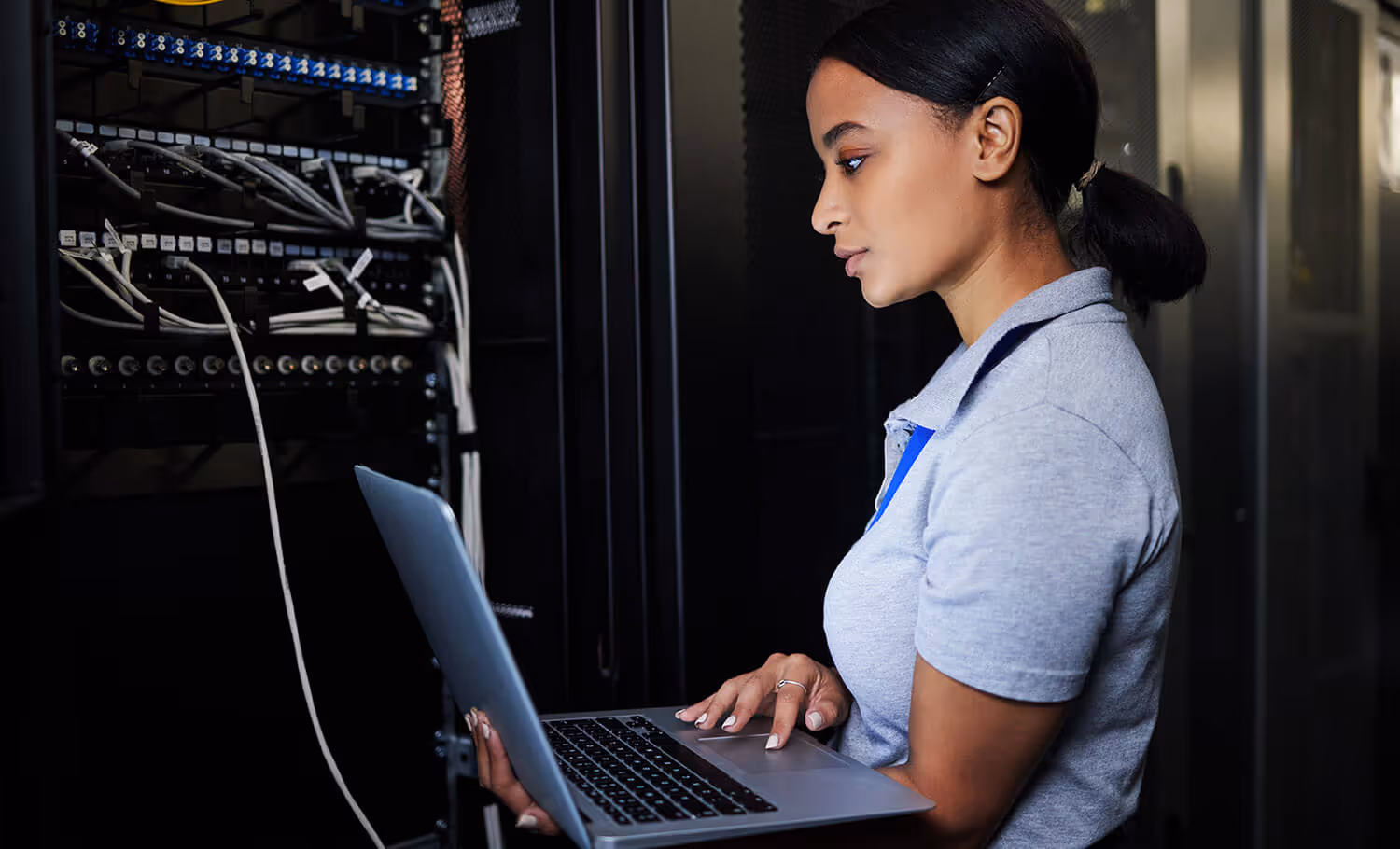 A person holding a laptop stands in front of a server rack with multiple connected cables.