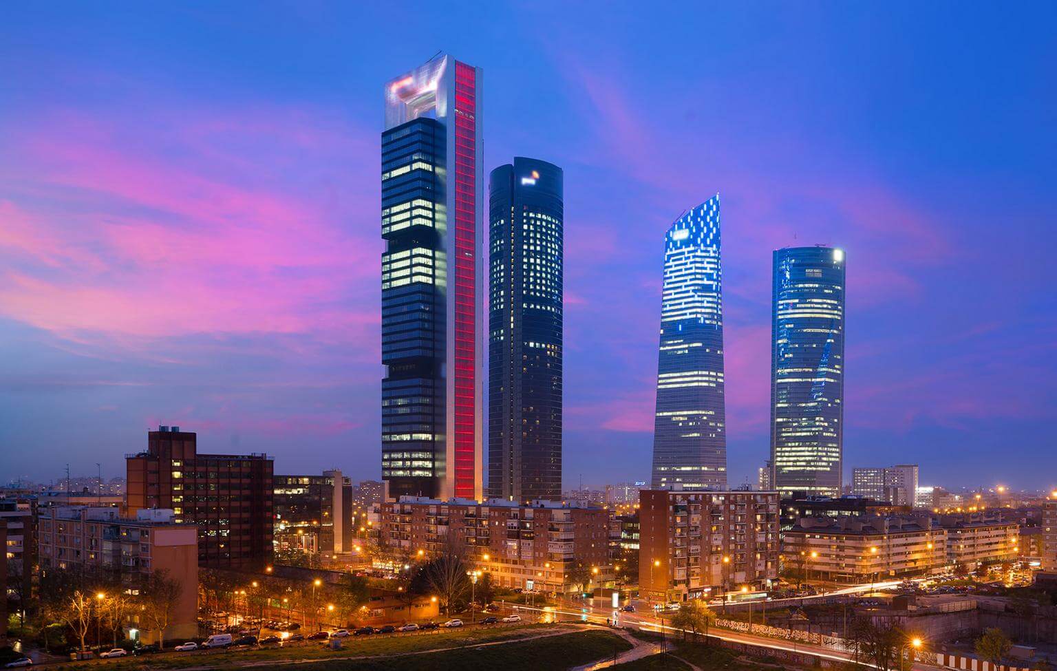Four tall illuminated skyscrapers rise above a cityscape at dusk with a pink and blue sky in the background.