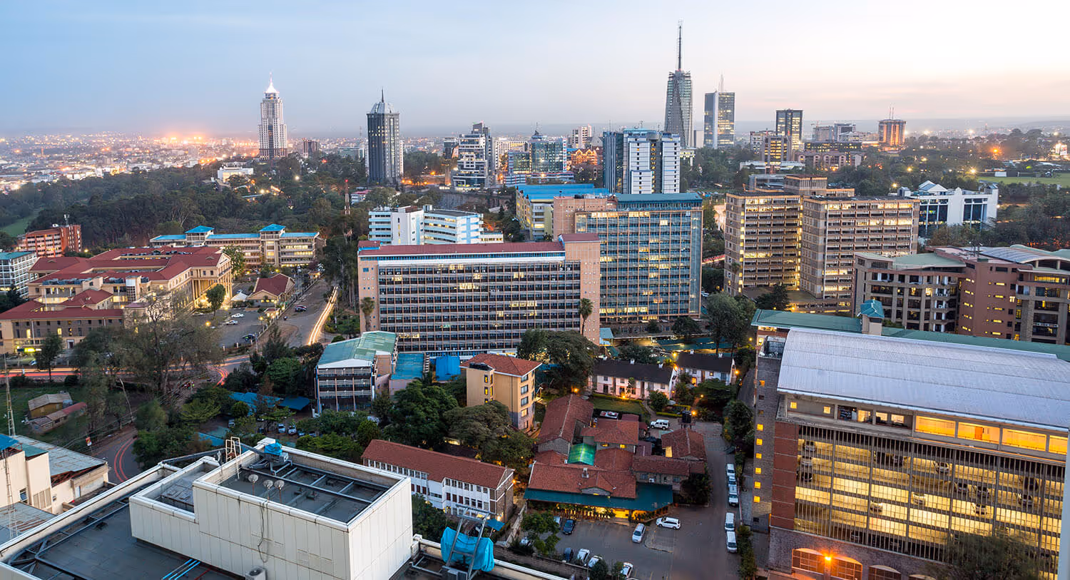 Cityscape with numerous multi-story buildings, trees, and lights at dusk under a clear sky.