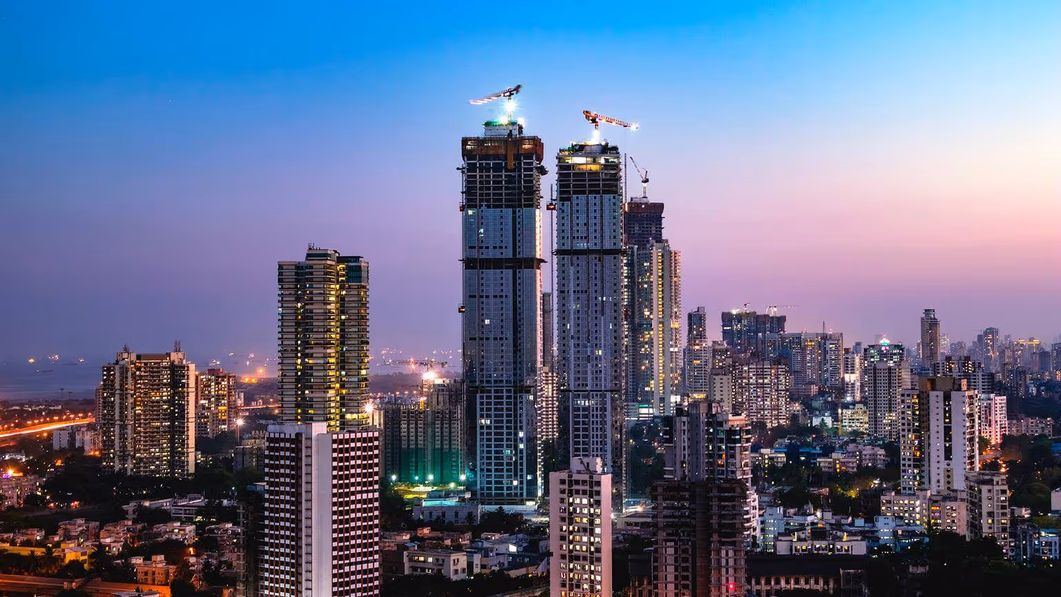 A cityscape at dusk with tall buildings, some under construction with cranes, and many windows illuminated.