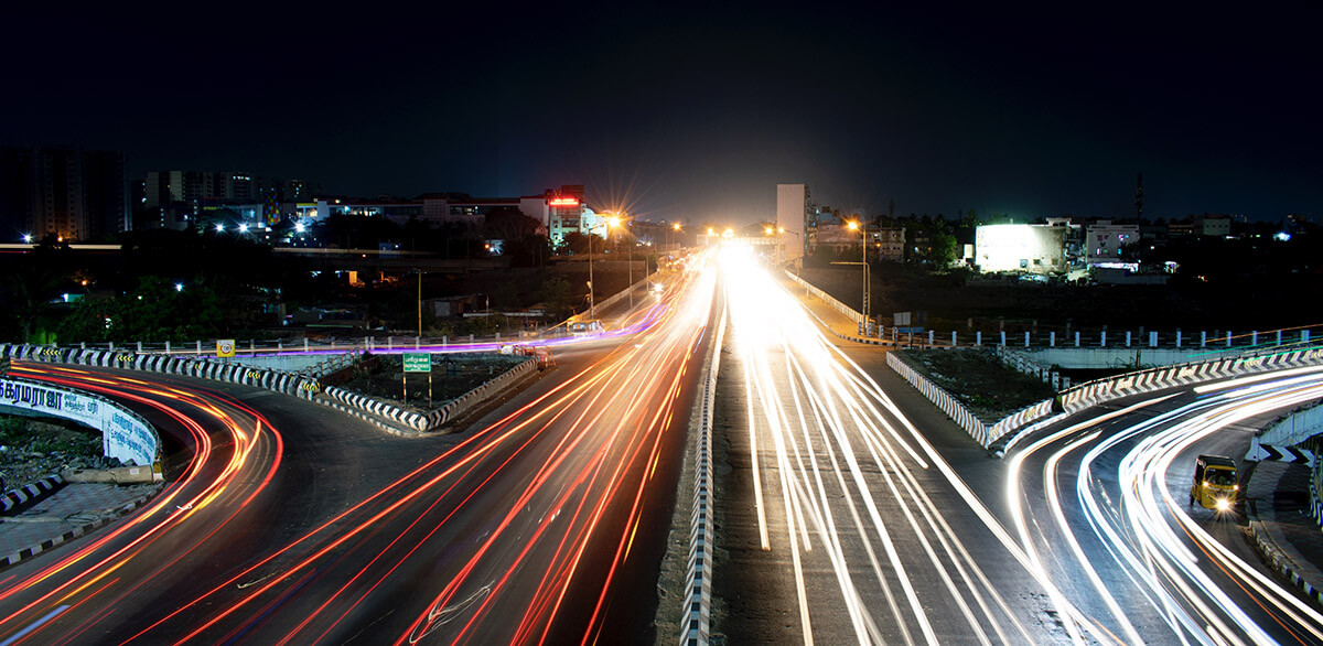 Long exposure photo of a divided highway at night with light trails from moving vehicles and buildings in the background.