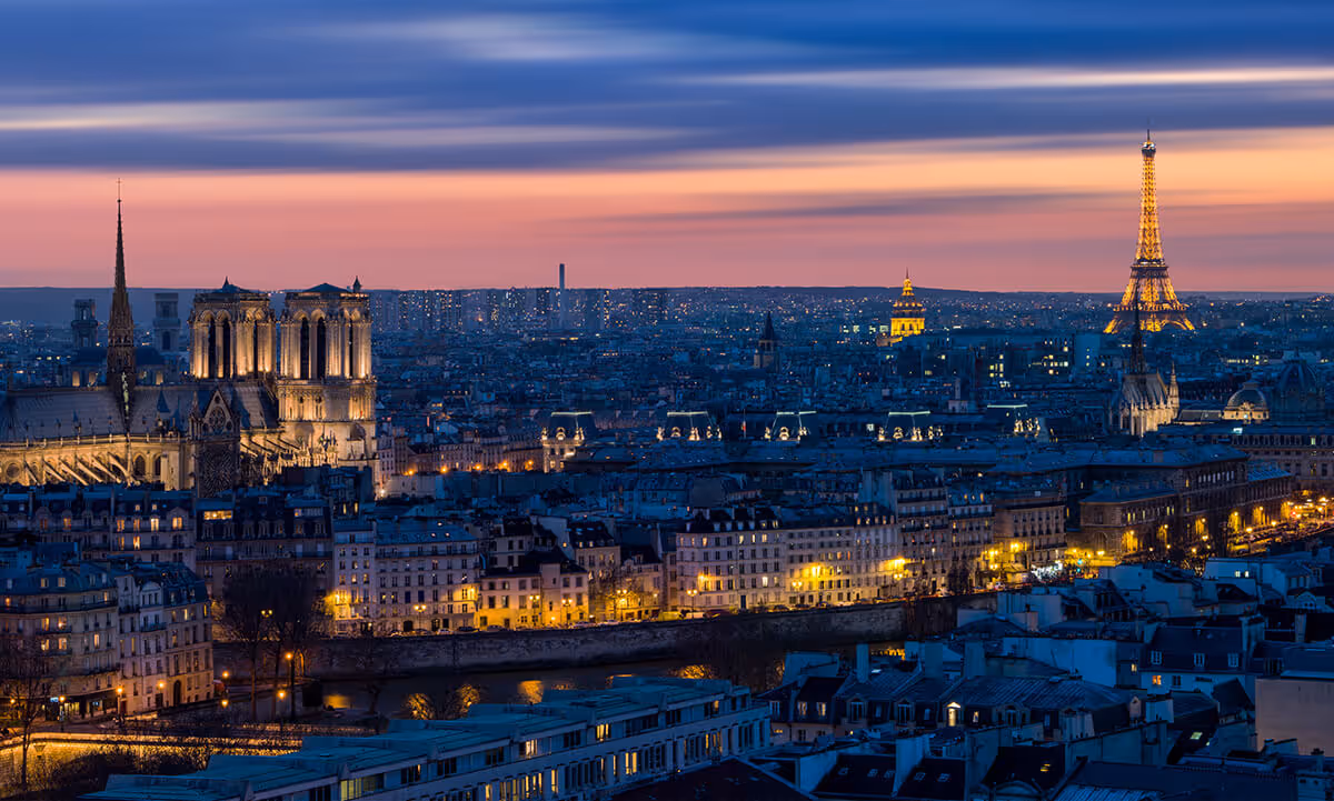 Cityscape at dusk with illuminated historic buildings, a river, and a prominent tower against a colorful sky.