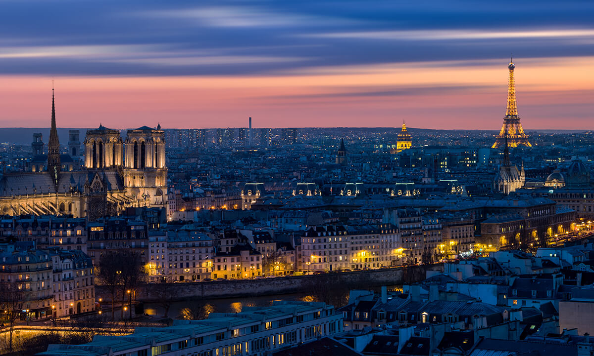 Cityscape at dusk with illuminated historic buildings, a river, and a prominent tower against a colorful sky.