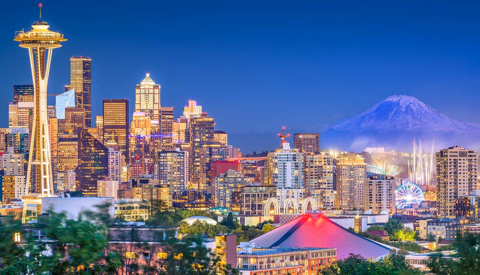 A city skyline at dusk with illuminated buildings, a distinct tower, and a snow-capped mountain in the background.