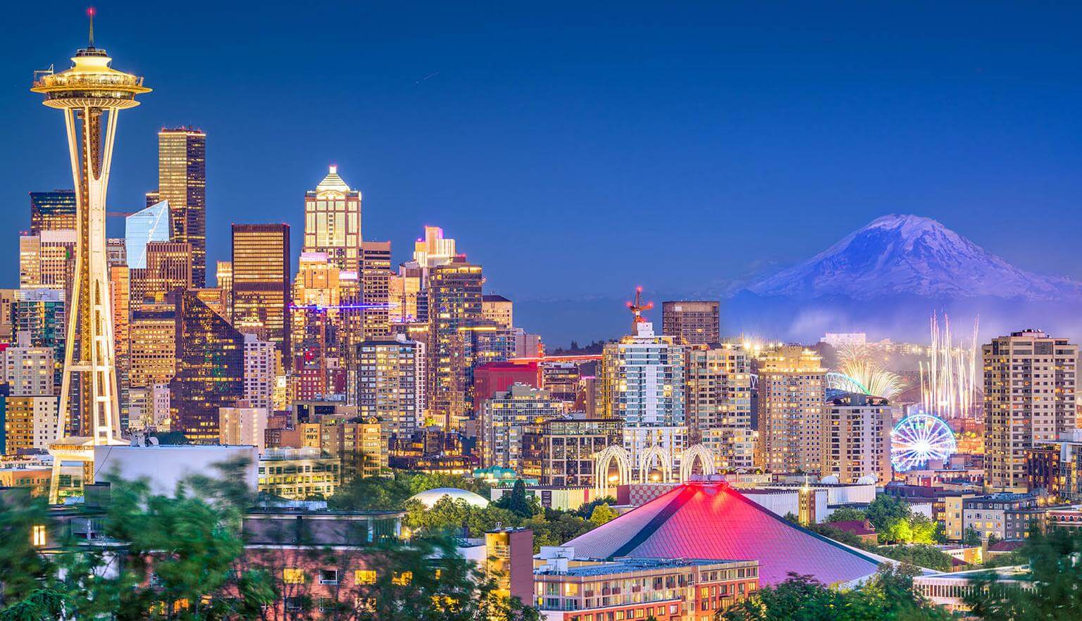A city skyline at dusk with illuminated buildings, a distinct tower, and a snow-capped mountain in the background.