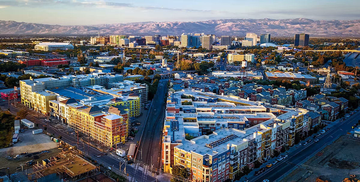 Aerial view of a city with colorful mid-rise buildings, train tracks, and mountains in the background.