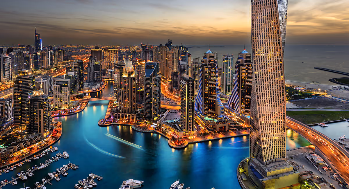 Tall modern skyscrapers surround a winding marina filled with boats at dusk, with city lights reflecting on the water.