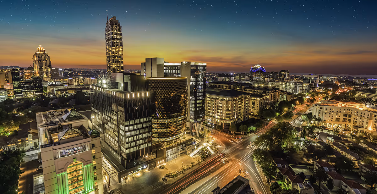 Cityscape with modern high-rise buildings illuminated at dusk, with light trails from traffic below and a starry sky above.