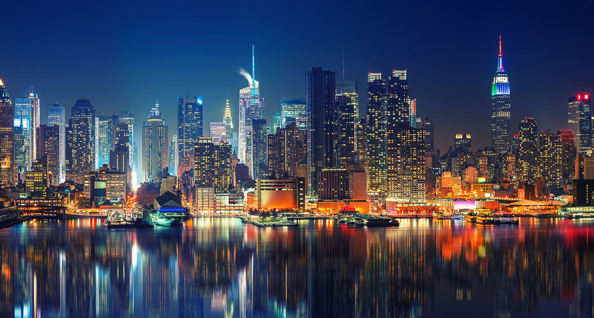 Tall buildings with illuminated windows at night are reflected in the calm water in front of the city skyline.